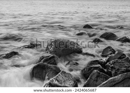 Captivating black and white photo of waves crashing onto black rocks, captured in soft focus. Foam forms a delicate white expanse resembling clouds. Taken at a beach in Lombok, Indonesia