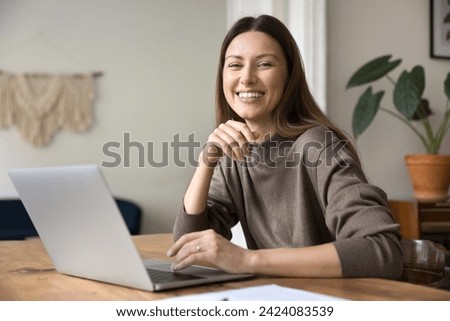 Happy successful entrepreneur woman working at laptop in home office, looking at camera for portrait with perfect toothy smile, posing at workplace table, enjoying remote job, online business