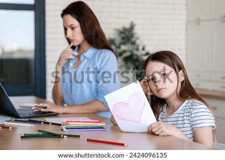 Bored child girl holding greeting card with heart to busy mother. Working parent having no time for kid, not paying attention, making urgent tasks.