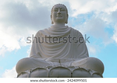 Horizontal picture of white statue of Buddha in Long Son Pagoda in Nha Trang, Vietnam, sitting on lotus by background of blue sky (view through green leaves of palm trees)
