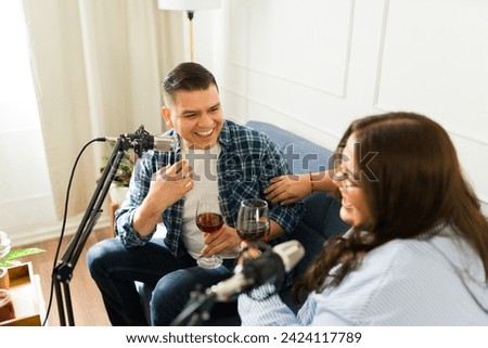 Attractive happy man laughing and having fun while talking with a microphone while recording a podcast with a woman guest drinking wine