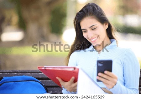 Happy student comparing phone and notes sitting in a park