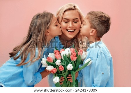 Happy Mother's Day! Cute charming girl and boy give their beautiful mother a bouquet of tulip flowers and kiss on the cheeks isolated on a pink background.