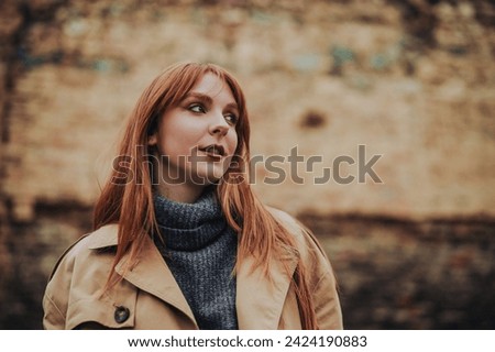 Fashion photo of a young ginger stylish woman standing on cold weather in warm outfit and looking away. Close up of a woman posing on a city street in old town whit rustic brick wall in a background.