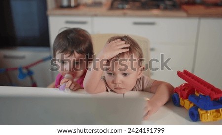 twin baby children playing laptop watching video in the kitchen. happy family kid dream concept. baby lifestyle twins playing video game on laptop looking at screen in kitchen