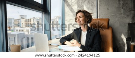 Portrait of successful lady boss, businesswoman in an office, working on her laptop, looking outside window, signing documents and papers.