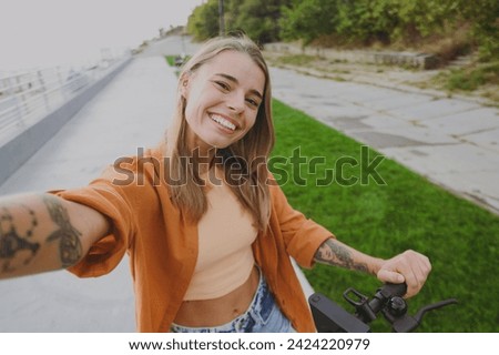 Close up young smiling woman wear orange shirt casual clothes sit on e-scooter do selfie shot pov on mobile cell phone walk rest relax in spring city park outdoors on nature. Urban lifestyle concept