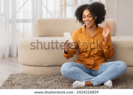 Joyful millennial African American woman wearing a mustard shirt and blue jeans sitting cross-legged on a rug, waving hand while video calling on her smartphone, enjoy video call