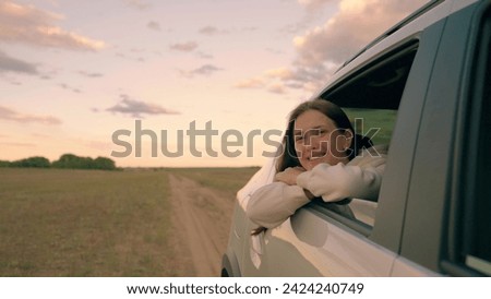 Beautiful girl travels smiling from car window, wind blowing her hair. Road trip on way to vacation. Young woman passenger looks out window, enjoys fresh breeze from car window, Happy look into future Royalty-Free Stock Photo #2424240749
