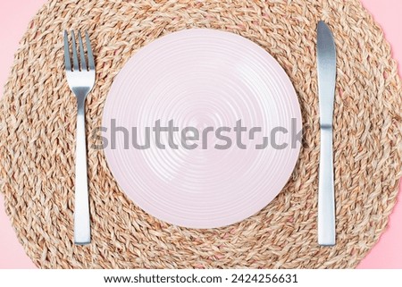 Empty pink plate, fork and knife against the background of wicker mat, intermittent fasting concept.