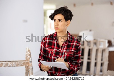 Focused interested dark-haired girl working on renovation and remodeling her house, making notes for space planning .. Royalty-Free Stock Photo #2424258713
