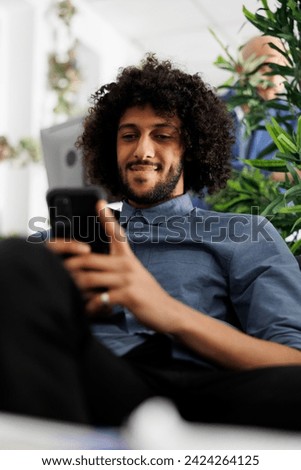 Smiling arab executive manager texting in social network on smartphone in business office. Start up company employee chatting on mobile phone while sitting on couch in coworking space