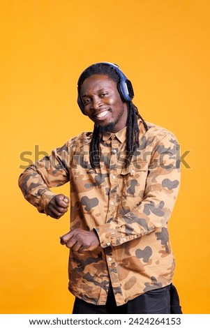 African american man listening music using headphones, dancing alone in studio over yellow background. Portrait of smiling young adult enjoying joyful playlist, having fun on song rhythm