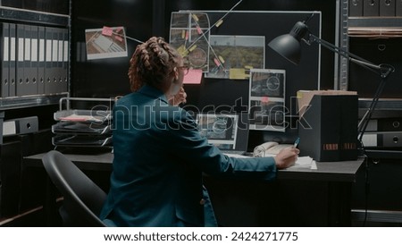 Police agent taking notes on case files in incident room, trying to solve criminal activity and catch suspect. Professional inspector examining research and surveillance photos. Royalty-Free Stock Photo #2424271775