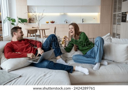 Laughing joyful wife holding smartphone showing funny short video, photo in social network to smiling husband. Cheerful man with laptop computer on knees working online as freelancer, watching film