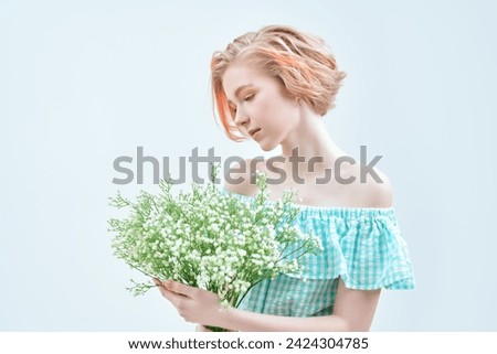 A cute blonde teenage girl with a short haircut in a summer sundress and a pearl necklace poses against a white wall with a bunch of flowers. Delicate spring-summer look. Beauty, cosmetics. Copy space