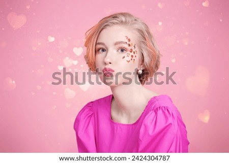 A cute blonde teenage girl with a short haircut poses in a pink dress with stickers in makeup and makes a funny face. Pink background with shining hearts. Spring-summer look.