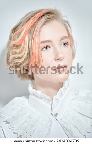 A cute teenage girl with a short haircut and blonde hair with orange streaks poses in a white blouse on a white studio background. Adolescence. Beauty, style and fashion.