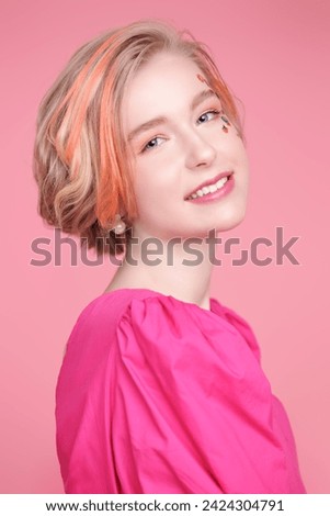 A lovely blonde teen girl with a short haircut with orange streaks poses with flower stickers on her face, wearing a pink dress. Pink background. Kids and teenage fashion. Gentle spring-summer look.