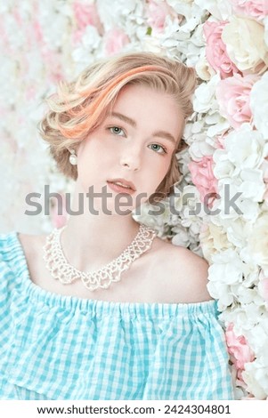 A cute blonde teenage girl with a short haircut in a summer sundress and a pearl necklace poses against a wall of white and pink flowers. Delicate spring-summer look. Beauty, cosmetics.