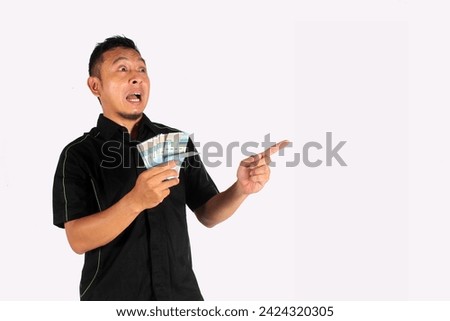 Attractive Asian man holding cash in one hand, with smile and surprised expression pointing at empty room and isolated white background
