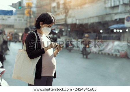 Asian pregnant woman wearing PM2.5 dust mask and using phone on the street. wearing mask protect against pollution, anti smog and viruses,  air pollution, and headache suffocating. City air pollution. Royalty-Free Stock Photo #2424341605