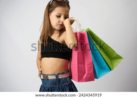 Portrait of a cute girl with colorful shopping bags. Little girl wearing summer clothes, standing, holding paper shopping bags on a white background with copy space.                        