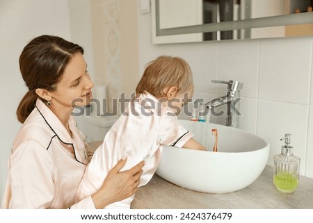 Mother watching daughter brushing teeth at home. Happy woman brushing teeth with her daughter. Little girl brushing teeth with her mom. Good morning concept. Daily routine and health care