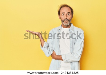 Caucasian middle-aged man in yellow studio impressed holding copy space on palm.