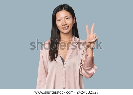 Young Chinese woman in studio setting joyful and carefree showing a peace symbol with fingers.