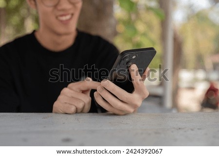 Close-up image of a happy young Asian man chatting with his friends on his smartphone while sitting at a table outdoor. people and technology concepts