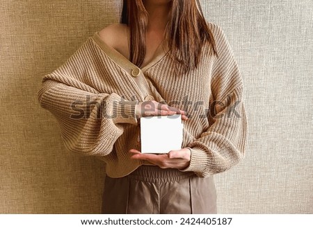 An unrecognizable brunette girl in a beige sweater holds a white square mockup in her hands. An empty sign in the hands of a girl.
