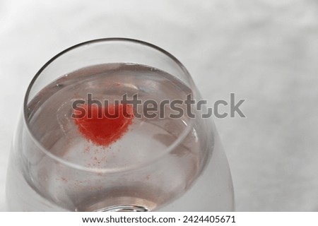 A glass is full of a clear liquid and a bright red, heart shaped ice cube floating in it Royalty-Free Stock Photo #2424405671