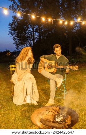 This image captures a serene evening scene where a man plays the guitar, serenading a woman seated next to him. They are both enjoying an intimate outdoor setting, illuminated by the warm glow of
