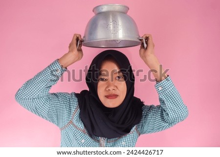 An Asian woman wearing a green shirt and black hijab is wearing a pot on her head. Cooking theme