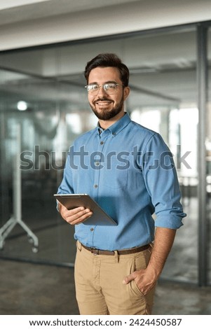 Smiling busy young latin business man company manager using tablet computer, happy hispanic businessman executive looking at camera holding tab working standing in office. Vertical portrait. Royalty-Free Stock Photo #2424450587