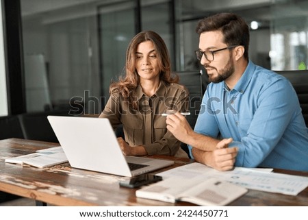 Two busy colleagues working together talking using laptop in office. Middle aged Latin female manager teaching young male worker looking at computer discussing business plan at office meeting. Royalty-Free Stock Photo #2424450771