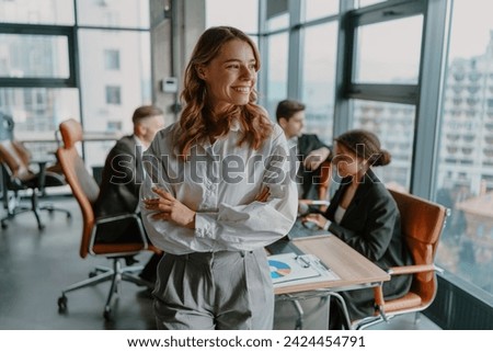 Smiling female entrepreneur with crossed hands standing in office on colleagues background