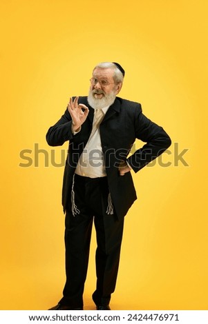 Portrait of smiling mature man in suit and black kippah making positive hand gesture against sunny yellow background. Purim festival, holiday, celebration Pesach or Passover, Judaism concept. Ad Royalty-Free Stock Photo #2424476971