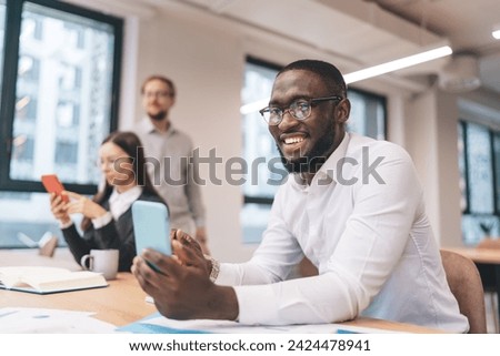 Cheerful young African American male employee in white shirt and eyeglasses smiling at camera while messaging via smartphone during workday with colleagues in modern office