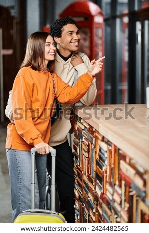 happy diverse couple with luggage laughing, woman pointing with finger at hostel reception desk Royalty-Free Stock Photo #2424482565