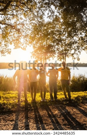 This image exudes the warmth of family unity with a group standing side by side by the lake, backlit by the radiant evening sun. The group is cast in a silhouette created by the bright sunlight Royalty-Free Stock Photo #2424485635