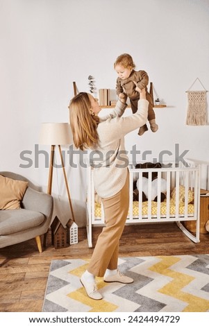 young woman holding little son while playing and having fun near crib in nursery room, motherhood
