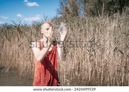 A man wearing a muscle shirt stands on the shore of a lake and has scooped water into the air. Royalty-Free Stock Photo #2424499539