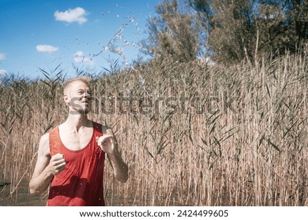 A man wearing a muscle shirt stands on the shore of a lake and has scooped water into the air. Royalty-Free Stock Photo #2424499605