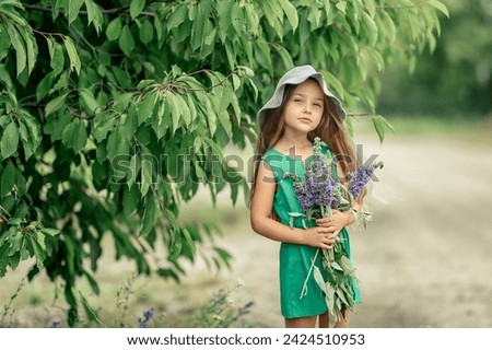 A girl in a hat with a bouquet of wildflowers stands on the road under a tree and looks at the camera. A little girl is walking with a bouquet of flowers in the park.