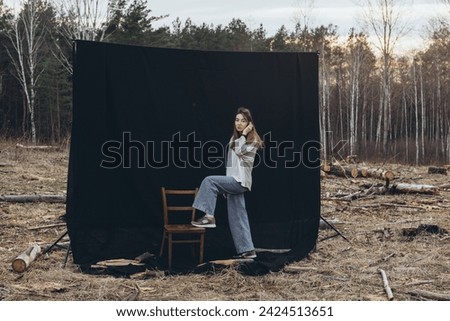 A beautiful sad girl sits on a chair, on a black background, set among deforestation at sunset. All around are tree stumps. Deforested forest. Destroyed nature