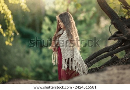 A beautiful little girl with long blond hair wrapped in a knitted blanket stands in the forest on a hill. A girl stands thoughtfully near a tree branch in the park. 