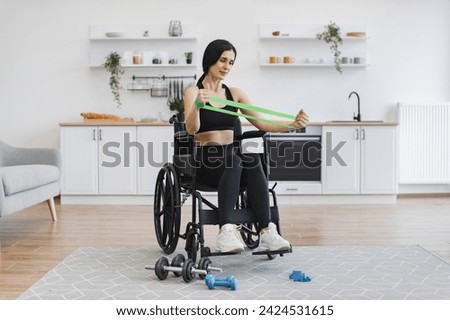 Positive woman in sportswear gaining health benefits from training. Full length view of Caucasian female with mobility impairment doing seated exercises with stretching bands in open-plan kitchen. Royalty-Free Stock Photo #2424531615