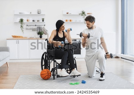 Rehab fitness for senior patients concept. Male physiotherapist or husband showing mature female patient in wheelchair how to exercise with dumbbells at home.